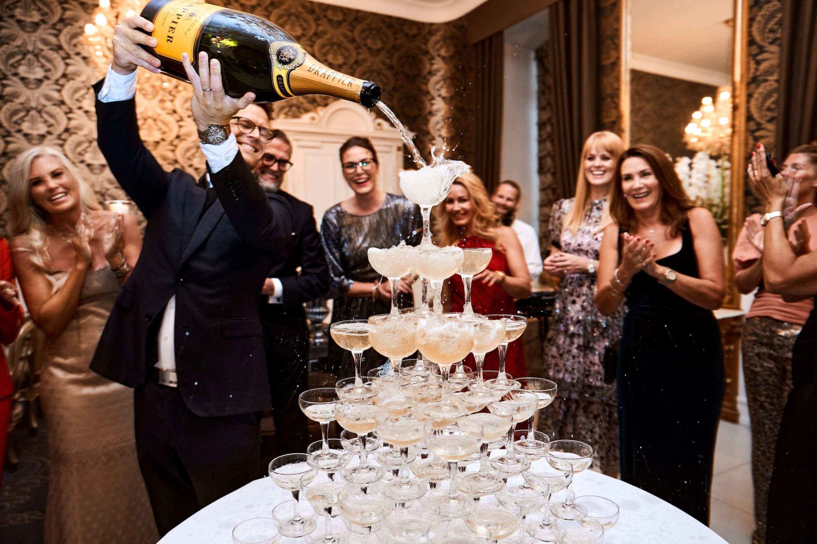 En mand hælder Champagne fra en kæmpe flaske udover et bjerg af champagne-glas som står oven på hinanden