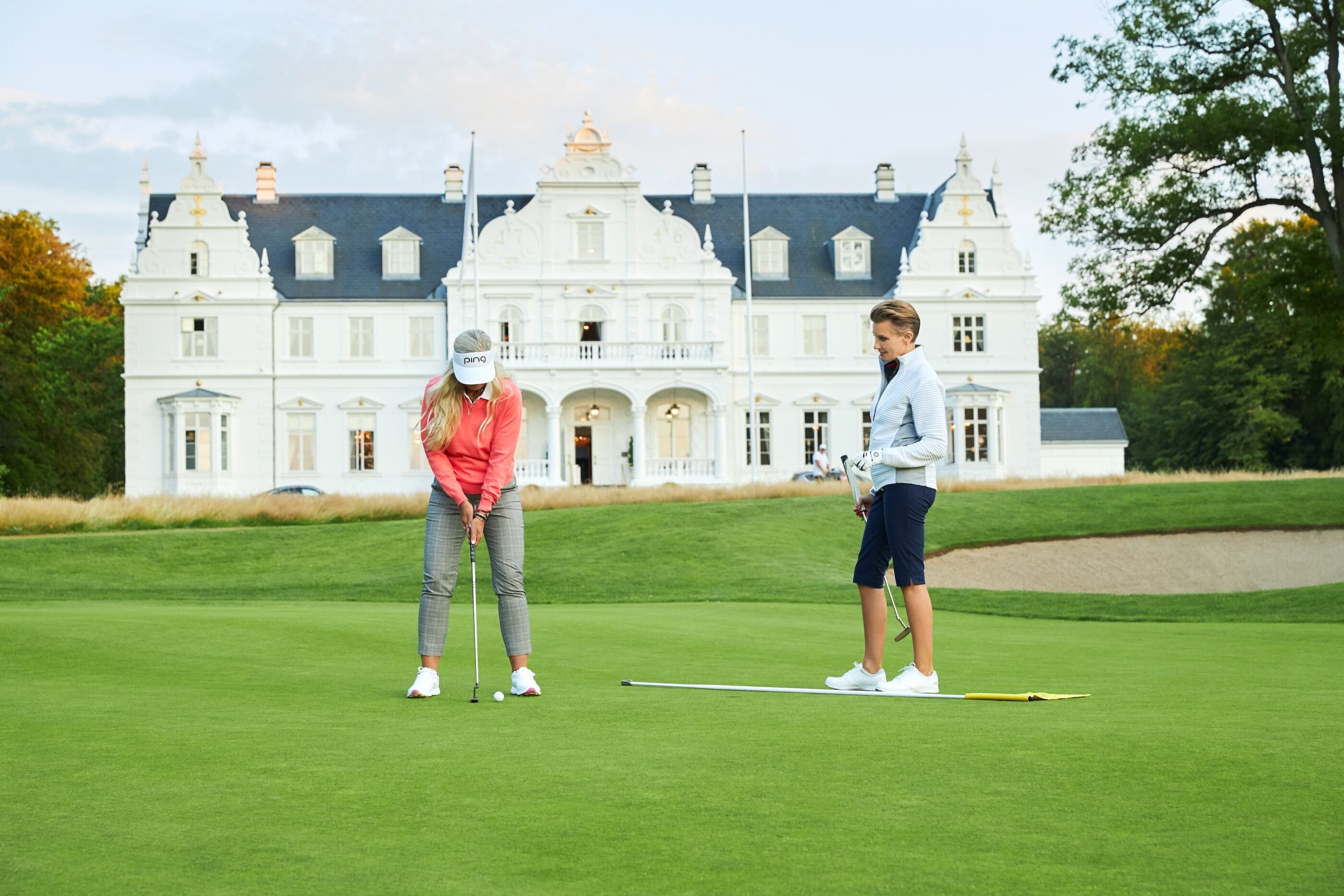 En mand og en kvinde iført golf tøj står på golfbanen med hotellet i baggrunden og kvinden skal til at slå til en golfboldt med sin kølle